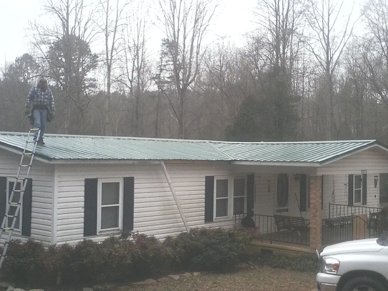 Worker on a ladder beside a new green metal roof on a ranch home in Hudson