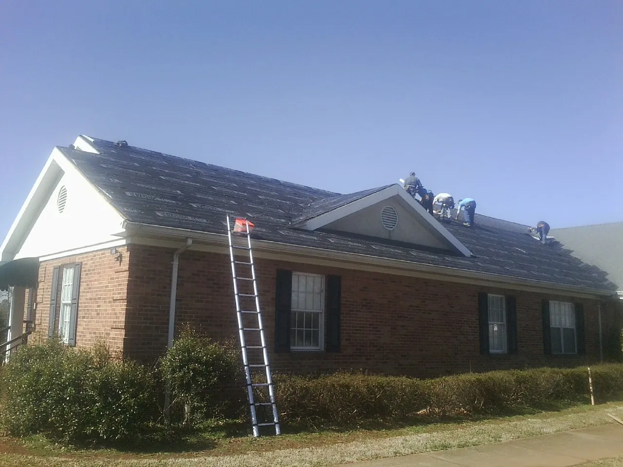 Crew tearing shingles off a brick ranch roof with a ladder set — Metal Roof Repair work by our crew in Hudson