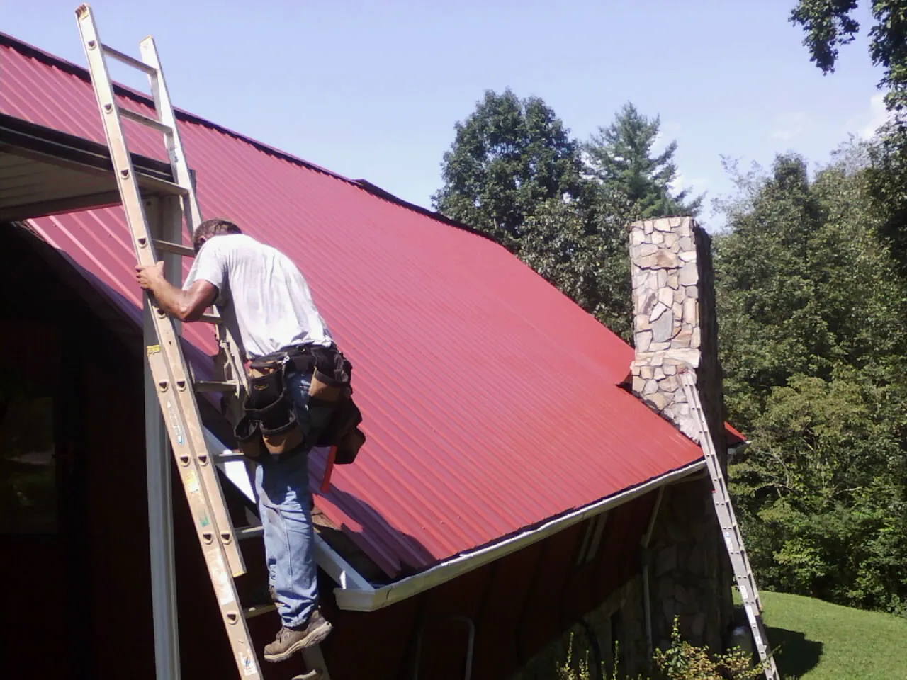 Roofer on a ladder beside a red metal roof and stone chimney — Soffit & Fascia Repair by Roofing Brothers in Hudson